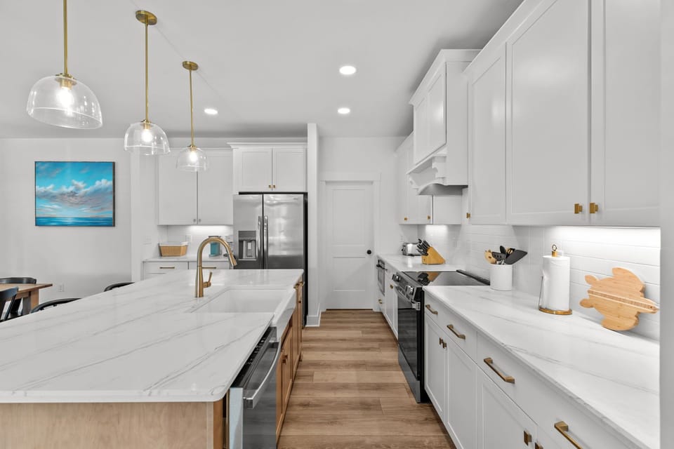 Bright white kitchen, marble island, gold faucet, glass pendants, black stools, stainless fridge, range hood, cabinets, wood floors.