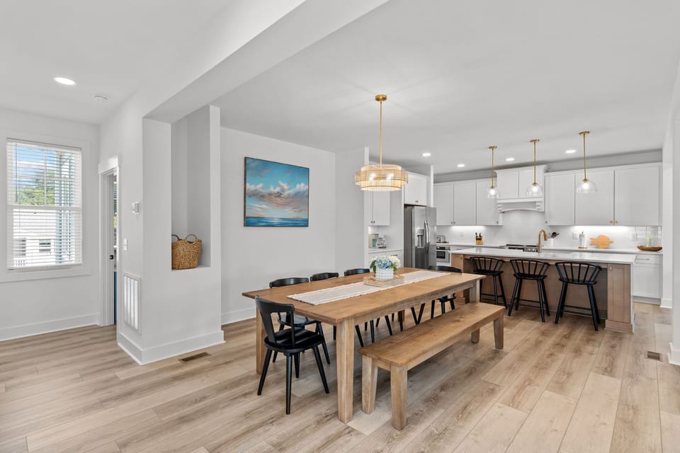 Bright dining room, long wood table, bench, black chairs, pendant light, white walls, doors, window, beach painting, wood floors.