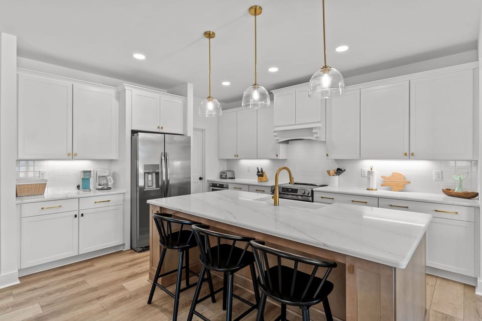 Bright white kitchen, marble island, gold faucet, glass pendants, black stools, stainless fridge, range hood, cabinets, wood floors.