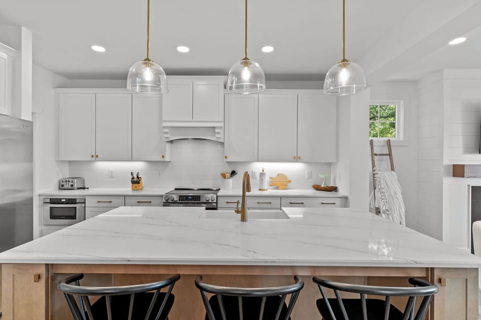 Bright white kitchen, marble island, gold faucet, glass pendants, black stools, stainless fridge, range hood, cabinets, wood floors.