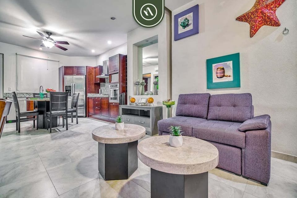 Relax in this vibrant, open-concept living area. It boasts a comfortable purple sofa, unique stone coffee tables, and flows seamlessly into the fully equipped kitchen and dining space.
