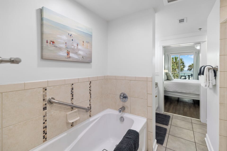 Slip into a soothing bath after a day by the water. Beige tiling and grab bars blend safety with comfort, while the bedroom beyond frames tropical palms and ocean breezes through the window.
