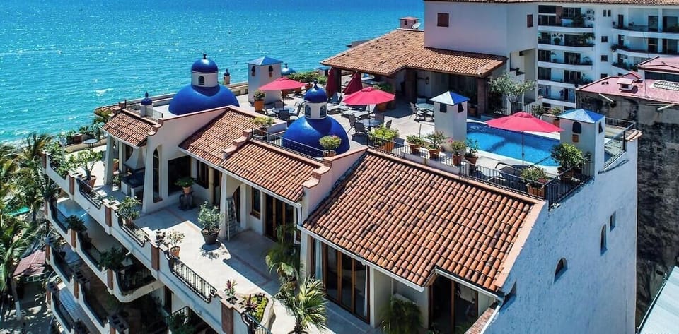 Panoramic drone perspective looking down over tile-roofed villas, blue dome architecture, and a central resort pool flanking the coastline.