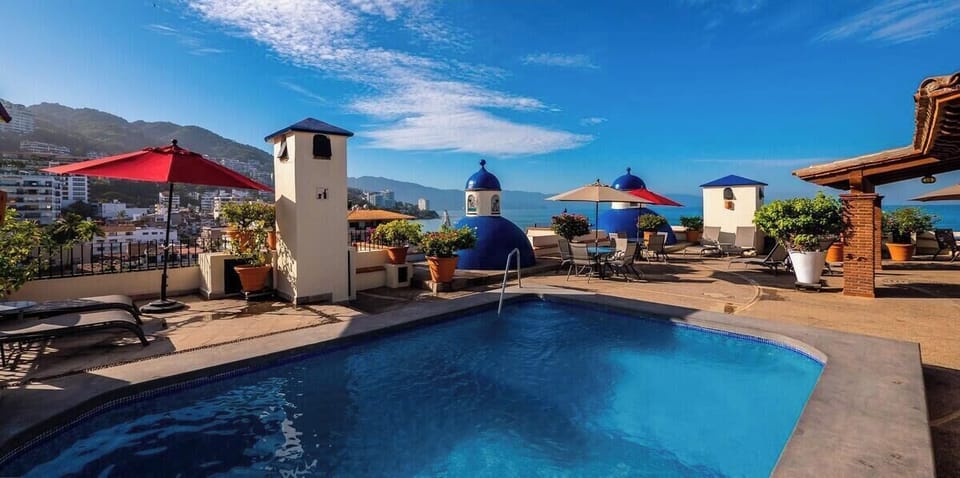 Rooftop swimming pool area framed by classic white towers, a signature blue architectural dome, and matching patio umbrellas.