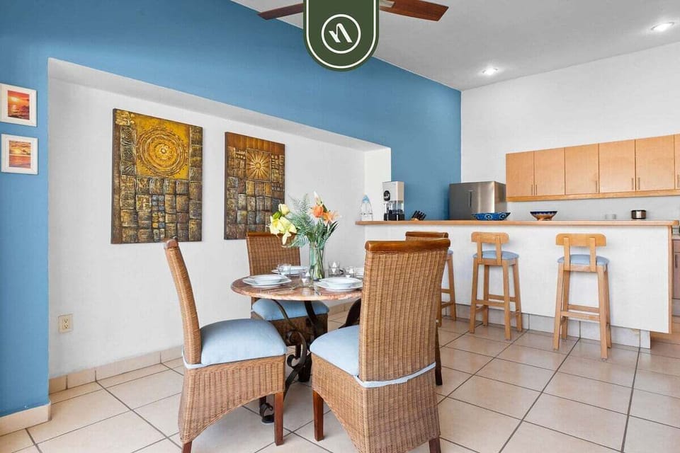 Round glass-top dining table paired with four woven wicker chairs, located adjacent to the kitchen breakfast counter.