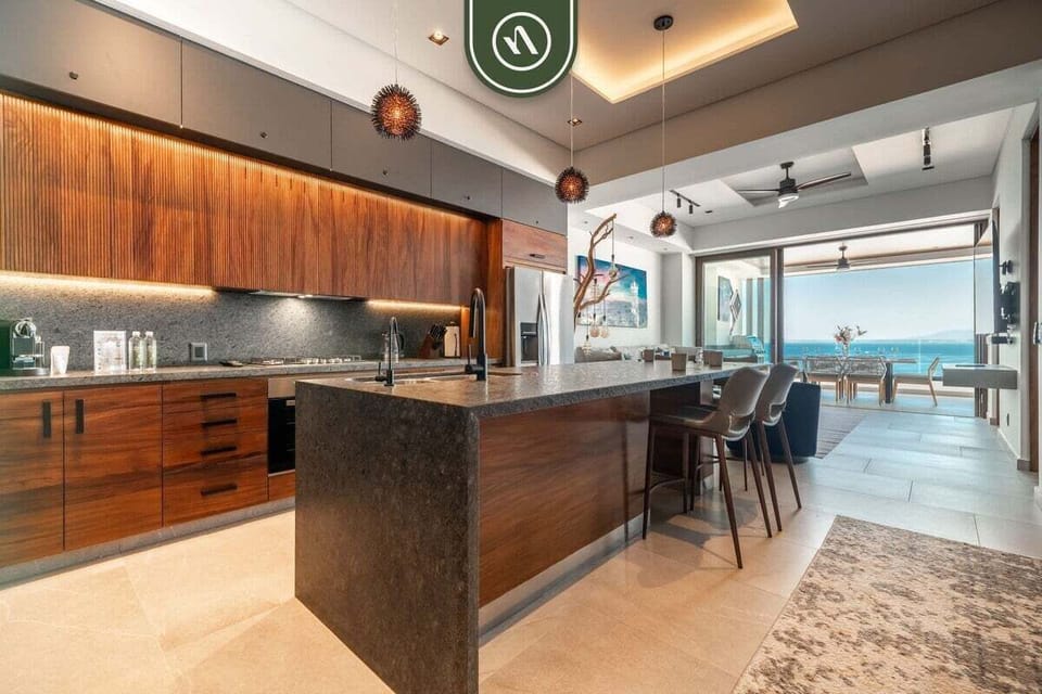Sleek gourmet kitchen showcasing a long granite breakfast island with bar stools, premium wood cabinetry, and a bright view toward the terrace.