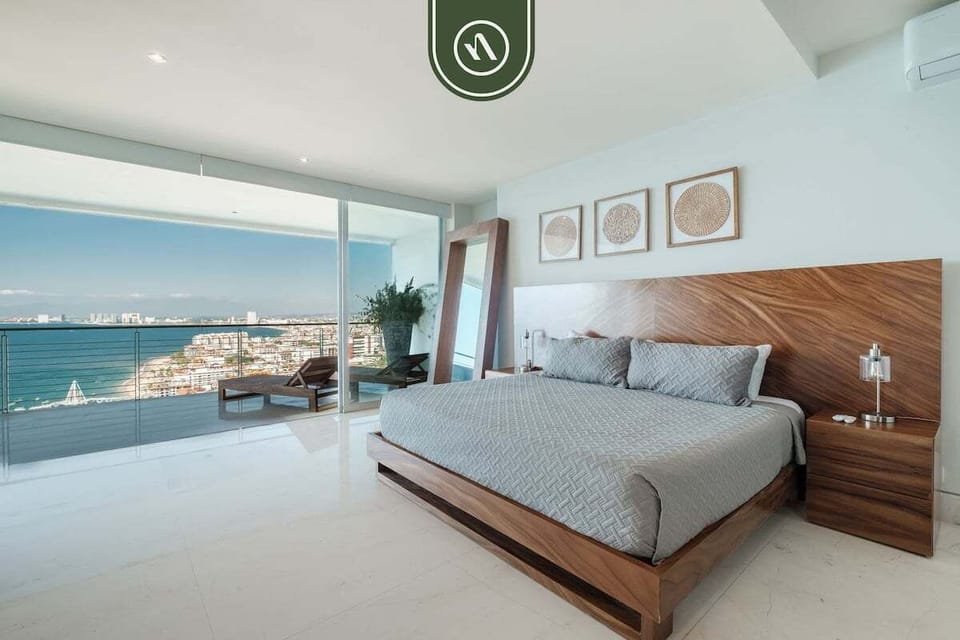 Elegant king bedroom with handcrafted wood furnishings, floor-to-ceiling glass doors, and direct views of the sparkling bay from bed.