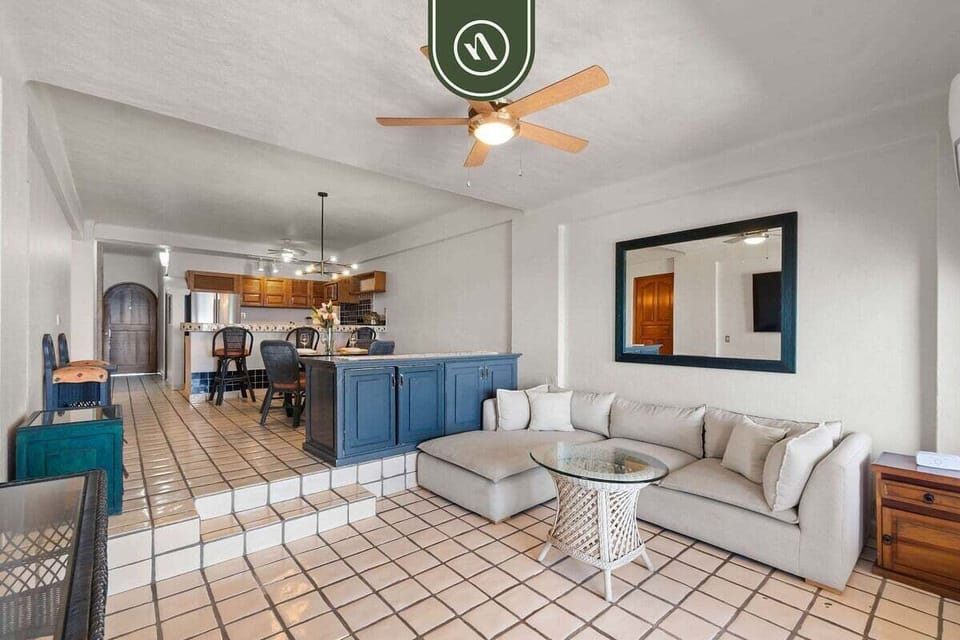 Tiled living space showcasing the large sectional sofa and glass coffee table, with steps leading up to the blue breakfast bar and kitchen.