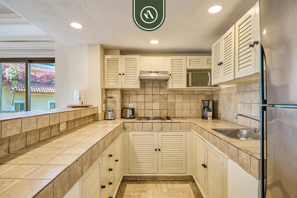 Functional U-shaped kitchen space showcasing beige tiled countertops, white shutter-style cabinetry, and essential cooking appliances.