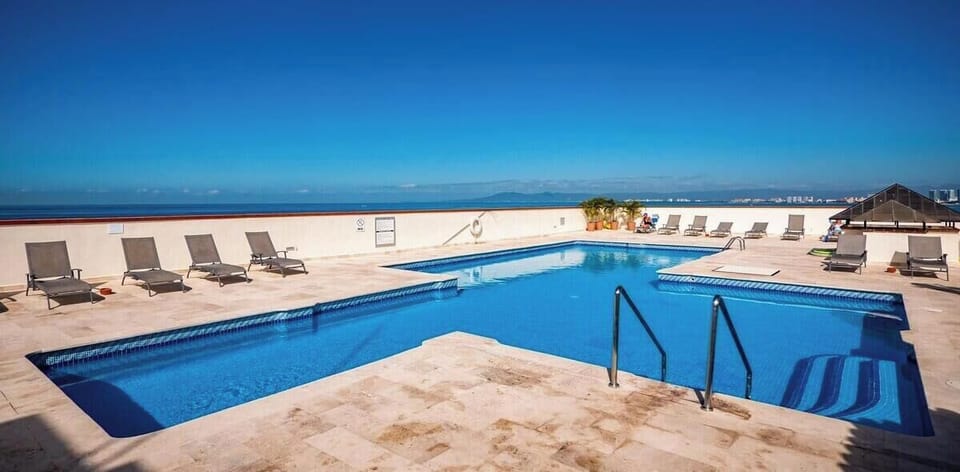 A sparkling rooftop swimming pool surrounded by a wide sun deck with lounge chairs under a clear blue sky.