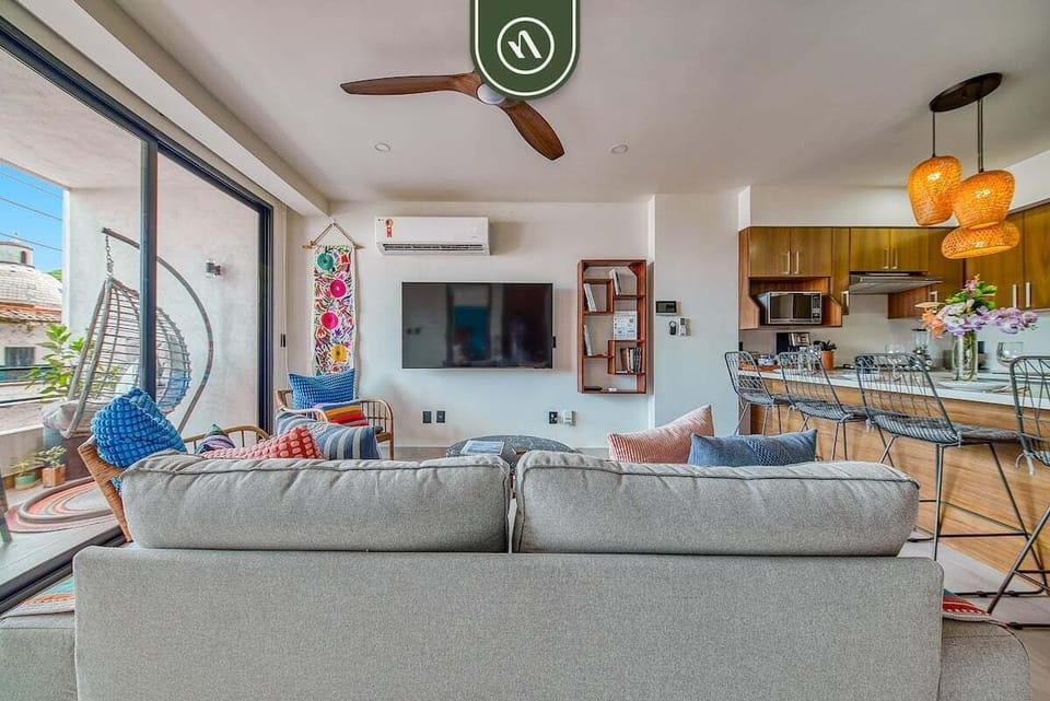 Relax in the spacious, open-concept living room. Viewed from the comfortable grey sofa, the area features a flat-screen TV, colorful textiles, and seamless access to the balcony and kitchen.