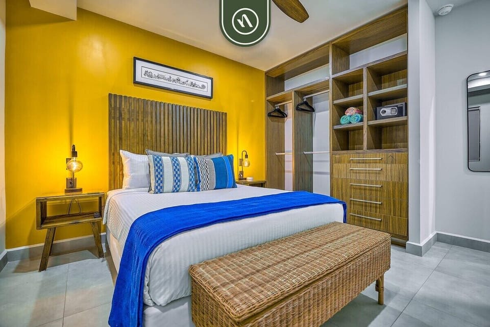 Settle into this bright and inviting bedroom. It features a sunny yellow accent wall, a plush bed with contrasting blue accents, a woven bench, and a spacious open closet.