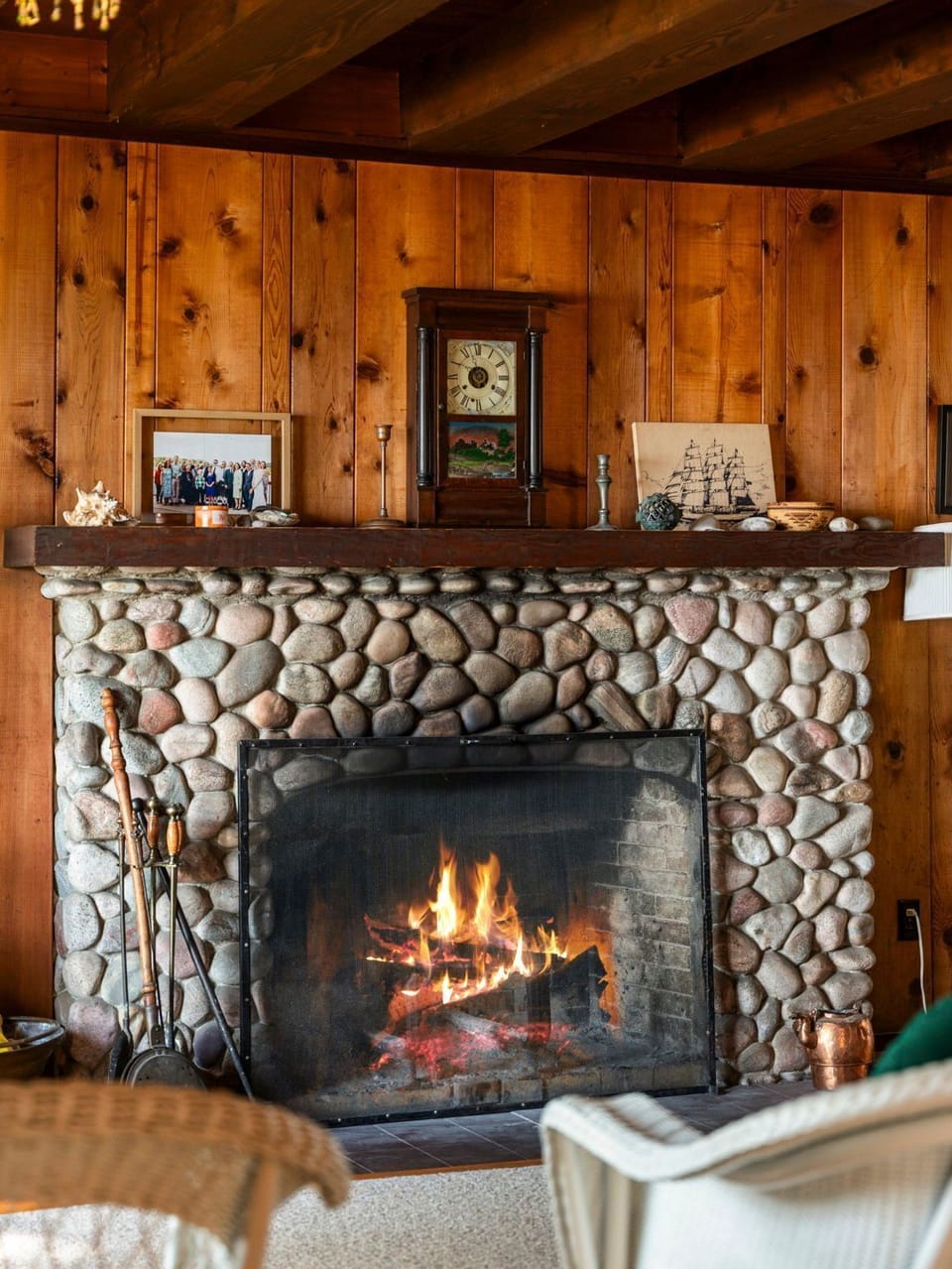 A cozy river stone fireplace with a lit fire and wood-paneled walls.