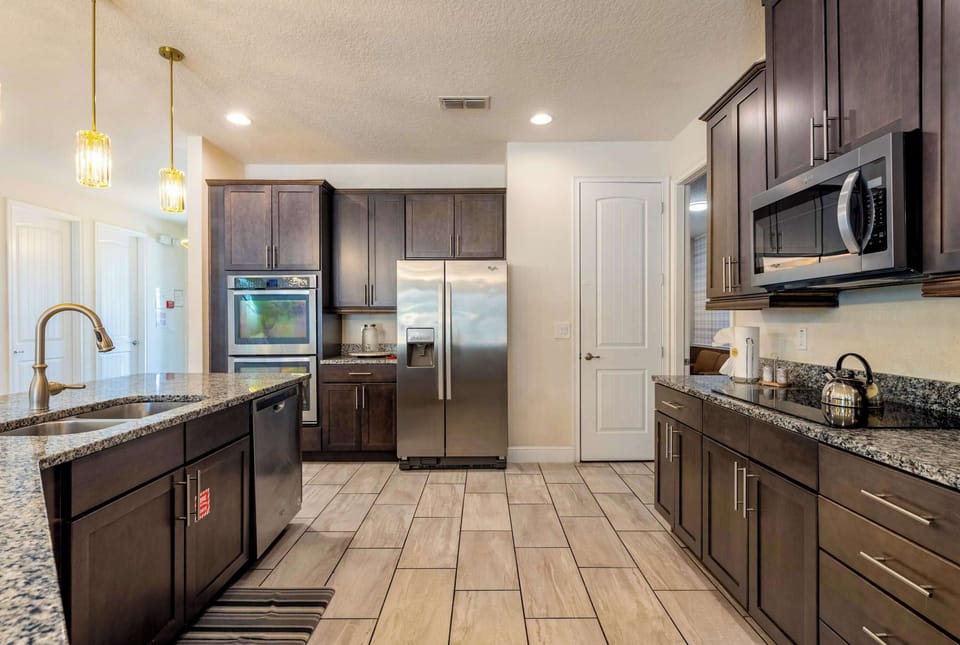 The chef's kitchen from the working side — double wall ovens, full-size stainless refrigeration, and granite prep counters that run the length of the room. Built for the catered dinner, the holiday spread, or the family breakfast for twenty.