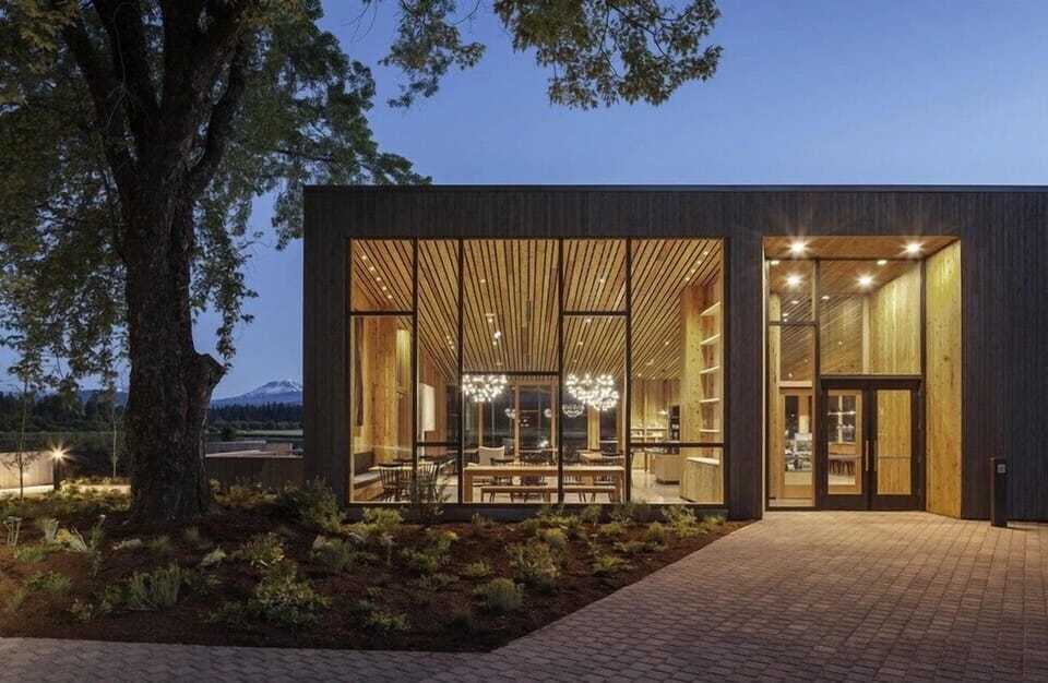 Modern architectural design showcases the Lakeside Bistro with warm wood interiors through floor-to-ceiling windows, nestled among mature trees with mountain views.
