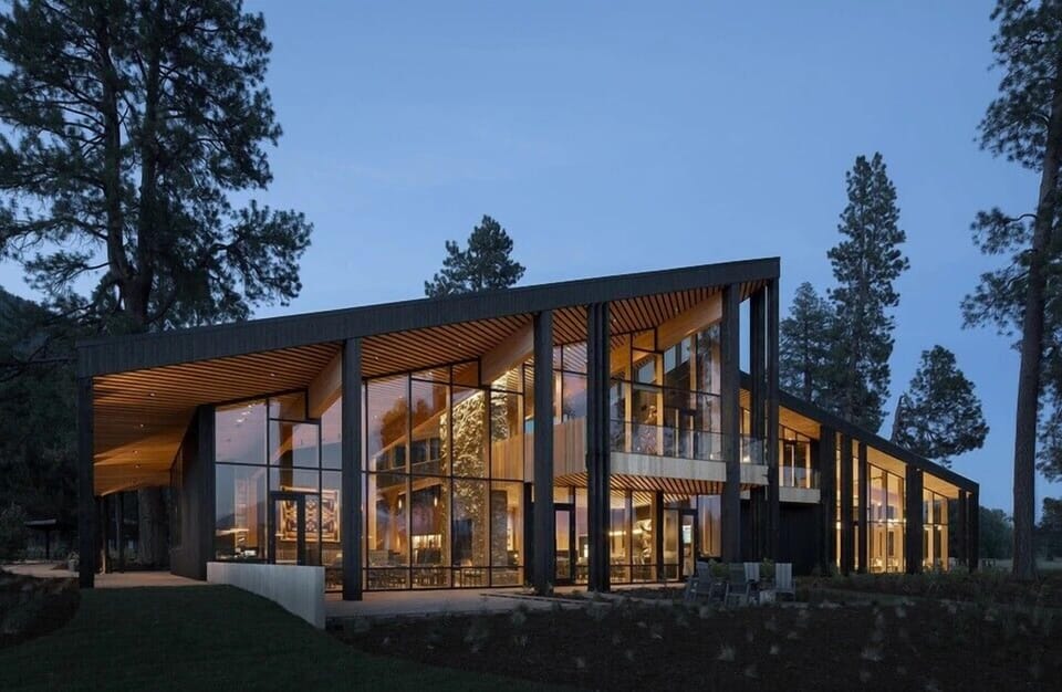 Stunning modern architecture showcasing the Lodge Restaurant nestled among towering trees with floor-to-ceiling windows with warm interior lighting at dusk.