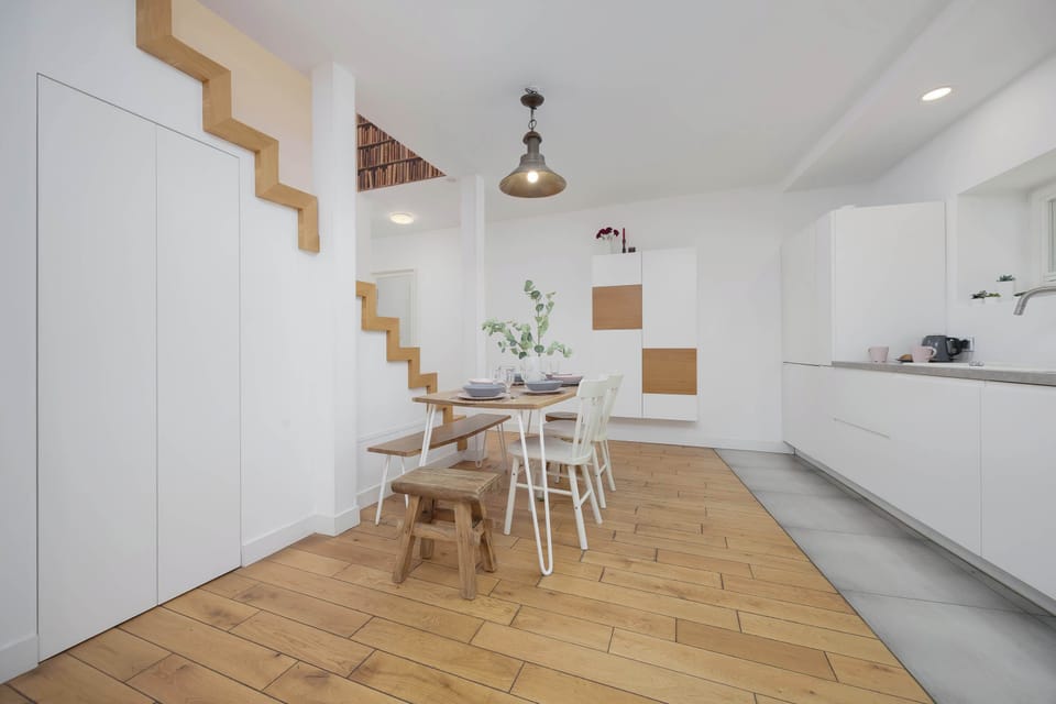 A bright dining area with a wooden table, minimalist kitchenette, and unique stairs leading.