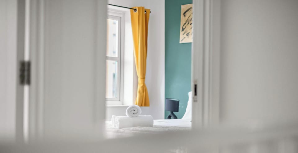 This perspective offers a charming glimpse into a bright, professionally styled bedroom. From the hallway, the eye is immediately drawn to the vibrant mustard yellow curtain and the teal accent wall, which create a beautiful.