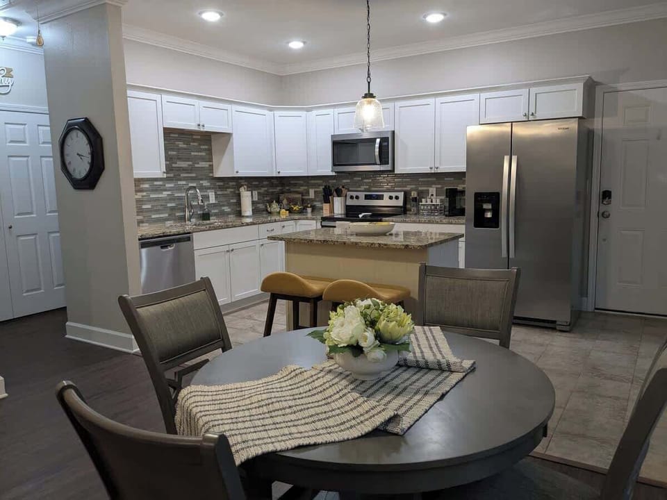 The dining table and kitchen as seen from the living room. The hallway to the left (with clock) shows the laundry closet and leads to the master bedroom. The back door is shown to the right of the refrigerator. 
