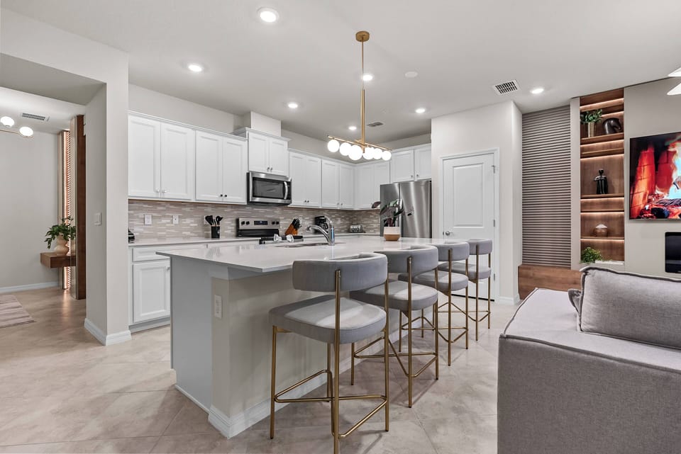 This stylish kitchen features sleek white cabinets and a spacious light stone island, great for meal prep or casual dining. Warm gold accents on the bar stools and modern lighting create an inviting space for gatherings.