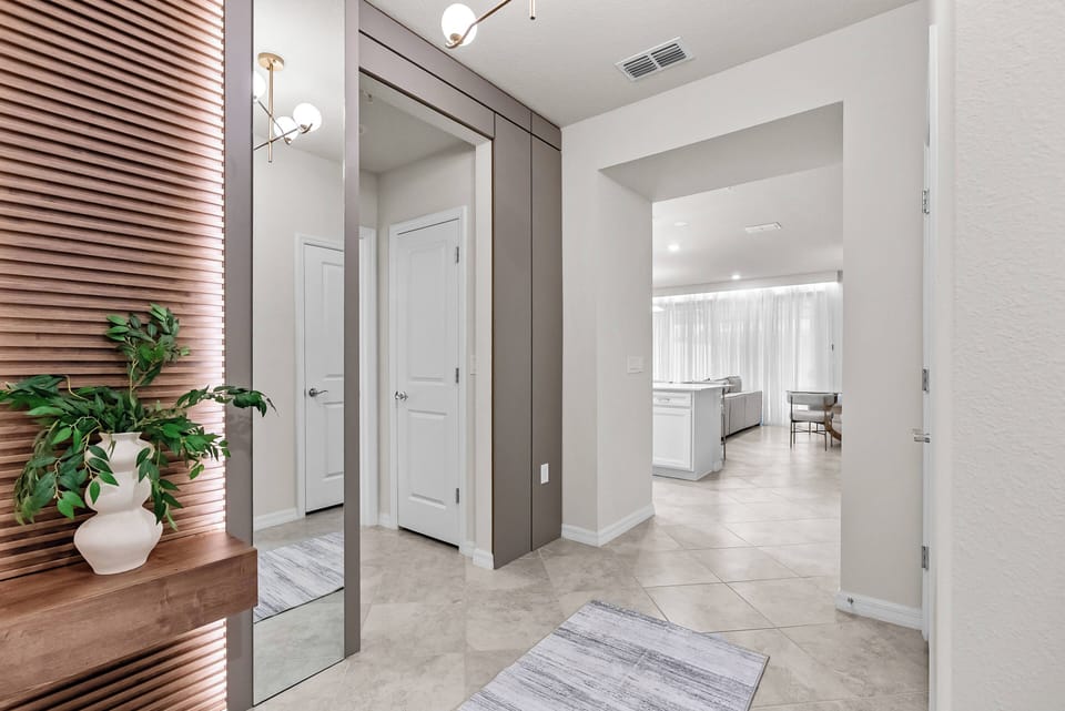 Enter a bright entryway with a sleek mirror and stylish wood accents that create a warm welcome. Soft, neutral walls and flooring lead you into an airy living area bathed in natural light from sheer curtains.