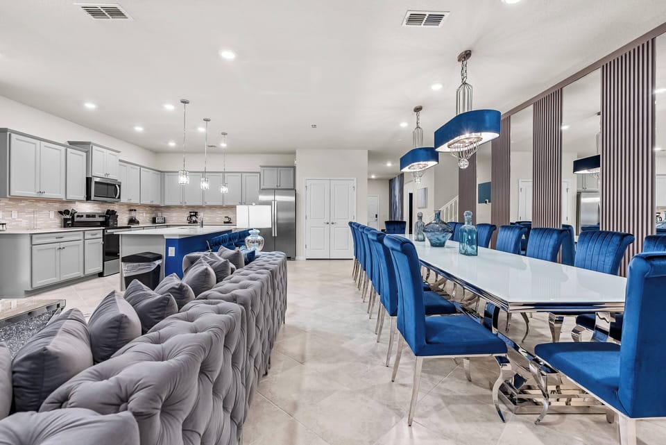 Experience a stylish dining area with a striking blue palette, plush chairs, and a sleek glass table, perfect for gatherings, seamlessly connected to a modern kitchen and beautifully lit by chic pendant lights for a vibrant.