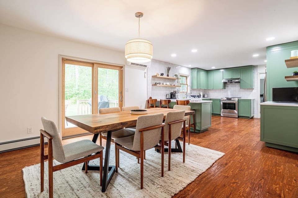 Bright and airy dining area with a large wooden table and modern chairs, seamlessly flowing into a fully equipped kitchen with stylish green cabinetry. Perfect for hosting meals and enjoying the view through sliding glass doors.