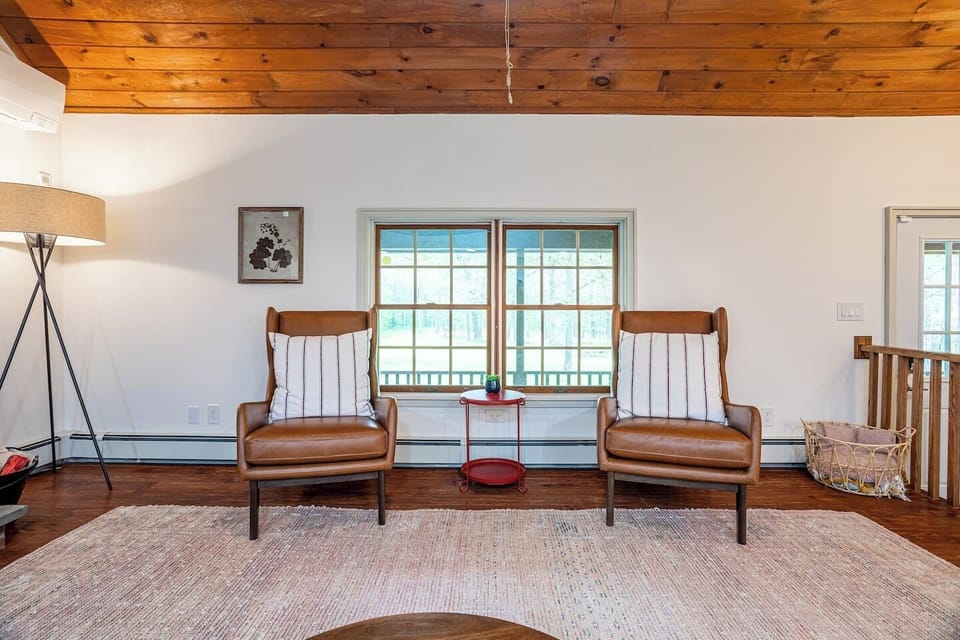 Charming seating nook in the living room, featuring two comfortable leather armchairs beneath a rustic wood ceiling. The large window offers views of the outdoors, making it an ideal spot for relaxing with a book.