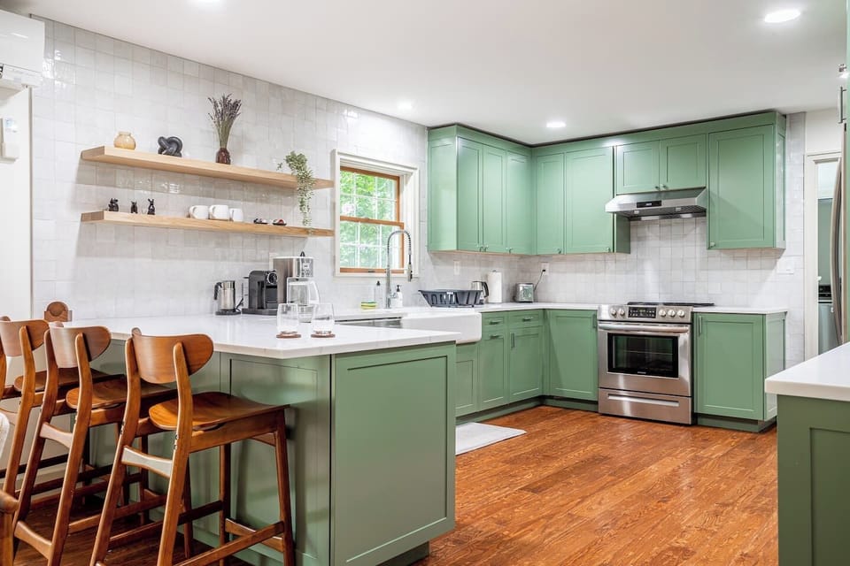Fully equipped kitchen with stylish sage green cabinetry, sleek white countertops, and a breakfast bar with wooden stools. Open shelving adds a modern touch, making it perfect for cooking and casual dining.