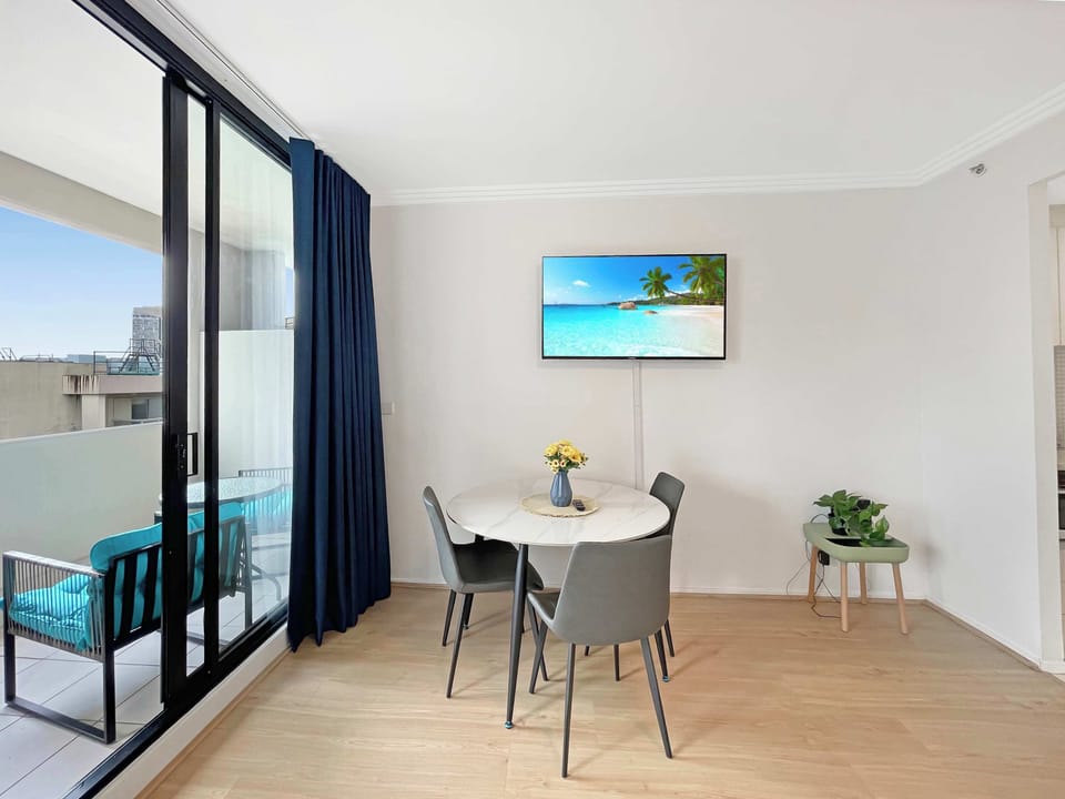 Open-plan bedroom and living space with queen bed, small dining area, and floor-to-ceiling windows with blue curtains overlooking city views.