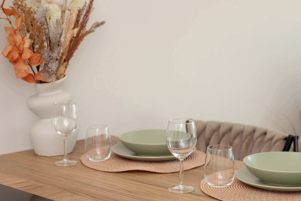 Close-up of a wooden dining table with green plates, glassware, and dried flowers in a vase.