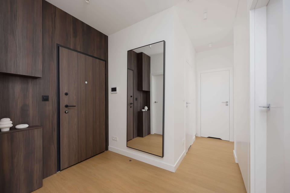 A hallway with a dark wooden front door, a wall mirror, and light wood flooring.