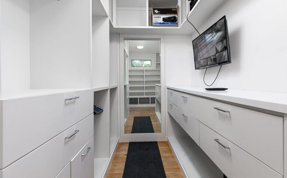 Walk-in closet with white built-in cabinetry, drawers, shelves, hanging space, a mounted TV displaying security camera feeds, and a mirror at the far end.