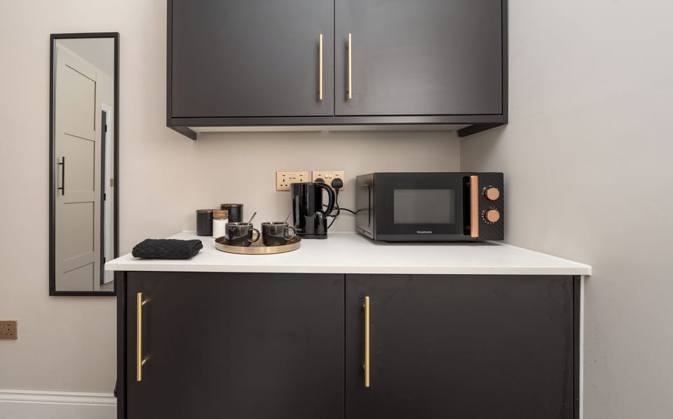 Compact yet practical, the kitchenette includes a two-seater dining space, microwave, toaster, kettle, fridge, and all essentials like tea, coffee, sugar, glasses, and cutlery. A wardrobe, wall mirror, iron, hairdryer, and board add extra convenience