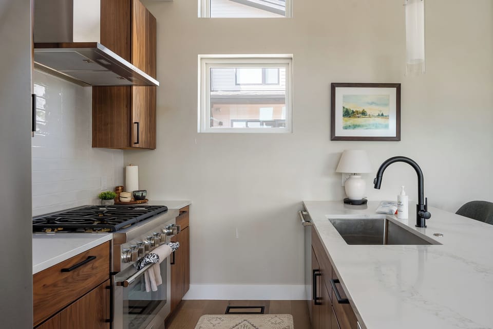 - Sleek sink with black faucet and wood cabinets   - Spacious counters ideal for meal prep and dining   - Window view adds charm to the modern kitchen