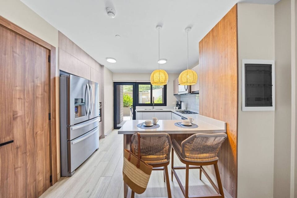 A modern kitchen with a breakfast bar, wicker chairs, pendant lights, and a sliding door opening to an outdoor space with a natural green view.