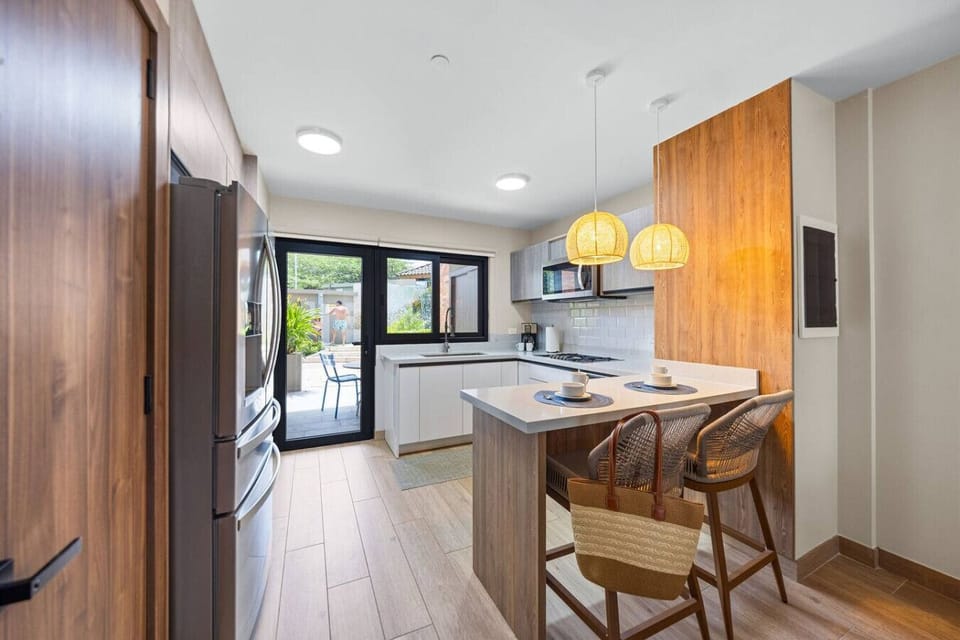 A modern kitchen with light wood and white cabinets, pendant lighting, a breakfast bar, and a sliding door opening to an outdoor area.