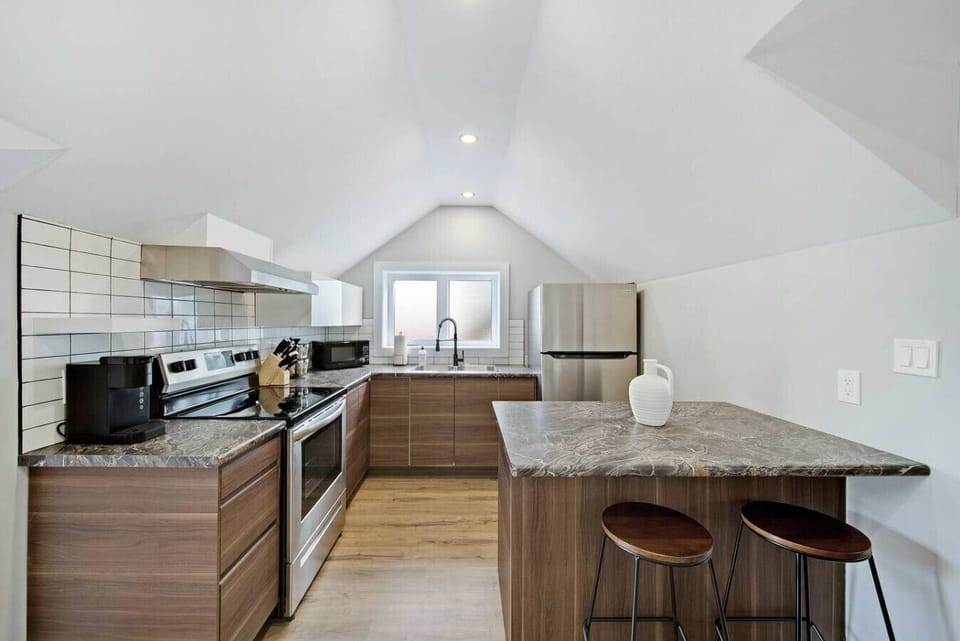 Modern full kitchen with stainless steel appliances, granite countertops, and a breakfast bar with two stools. Features white upper cabinets and sleek brown lower cabinets. Perfect for cooking and casual dining.