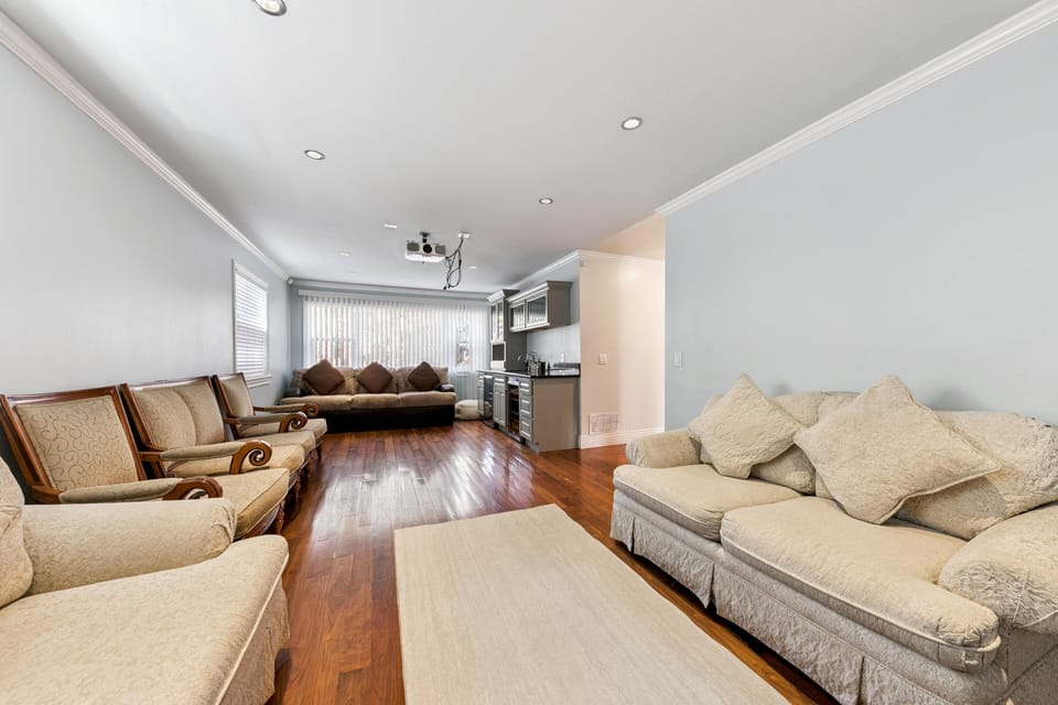 Long seating area with beige upholstered sofas and armchairs faces a wall-mounted projector above a kitchenette.