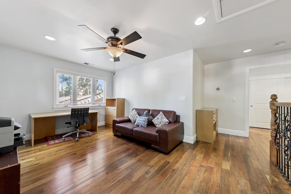 Upper-level room with hardwood floors, a brown leather loveseat, wooden desk, and ceiling fan with light.