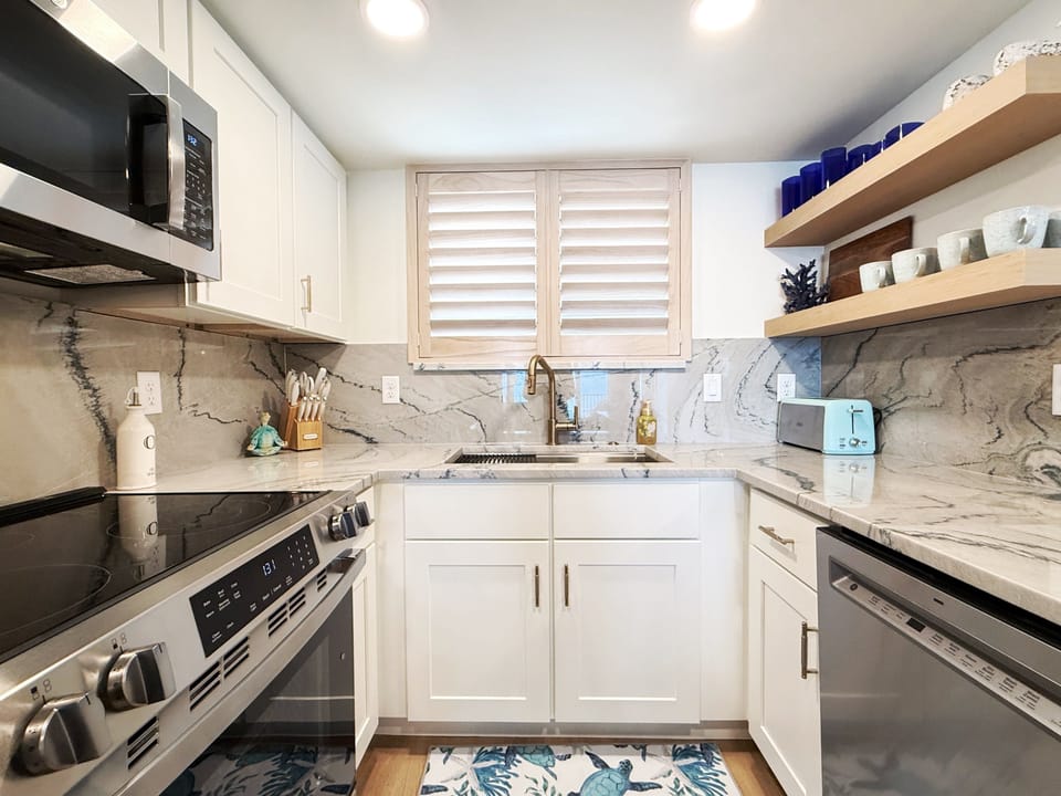 Cook with ease in this stylish kitchen featuring stainless steel appliances, marble countertops, and open shelving—perfect for preparing meals to enjoy after a relaxing beach day.