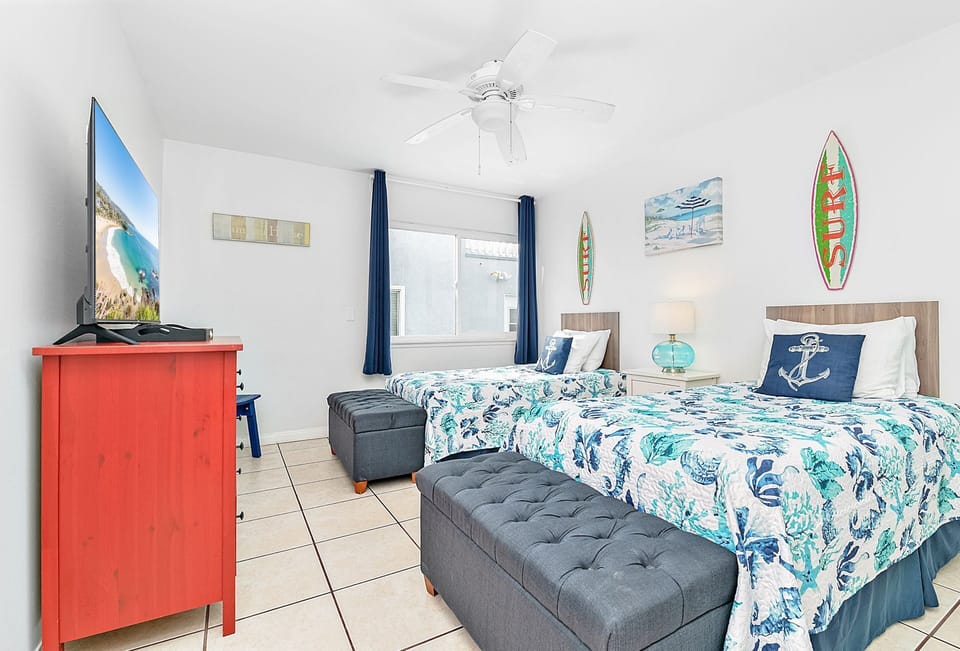 A bright, beach-inspired guest bedroom featuring two twin-size beds and a flatscreen TV
                