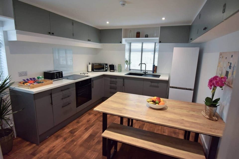 Modern, fully equipped kitchen with sleek grey cabinets, wood flooring, and a large dining bench. Includes oven, microwave, fridge-freezer, toaster, kettle and coffee machine. Bright, clean, and perfect for preparing meals or relaxing with a brew.