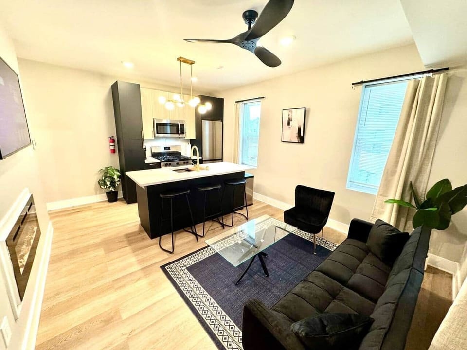 Modern kitchen featuring brand-new appliances, including a breakfast bar, dishwasher, microwave, and refrigerator. It seamlessly connects to the living room, which is furnished with a sofa bed, ana a large flat-screen TV.