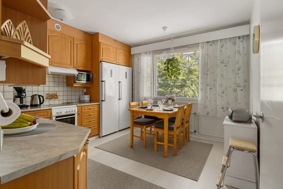 Bright and welcoming kitchen featuring warm wooden cabinetry and modern appliances including a double-door refrigerator, oven, microwave, and toaster. A cozy dining area with a wooden table and four chairs is set by the large window, allowing plenty of na