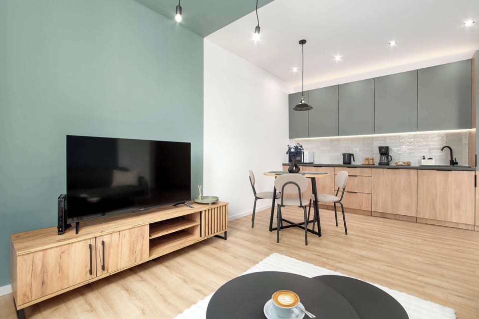 A living room with a sofa and a TV unit, connecting seamlessly to the kitchen. Large windows provide natural light, enhancing the airy feel.