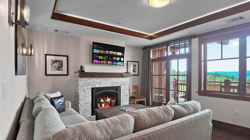 Sunlit living room with balcony, forest views, and crackling fire.
