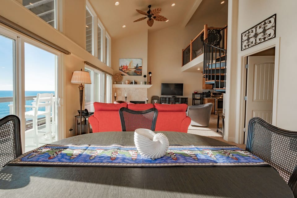 The Coastal Villa features a bright open-concept living area with vaulted ceilings, ocean-view windows, and a dining table set for four.