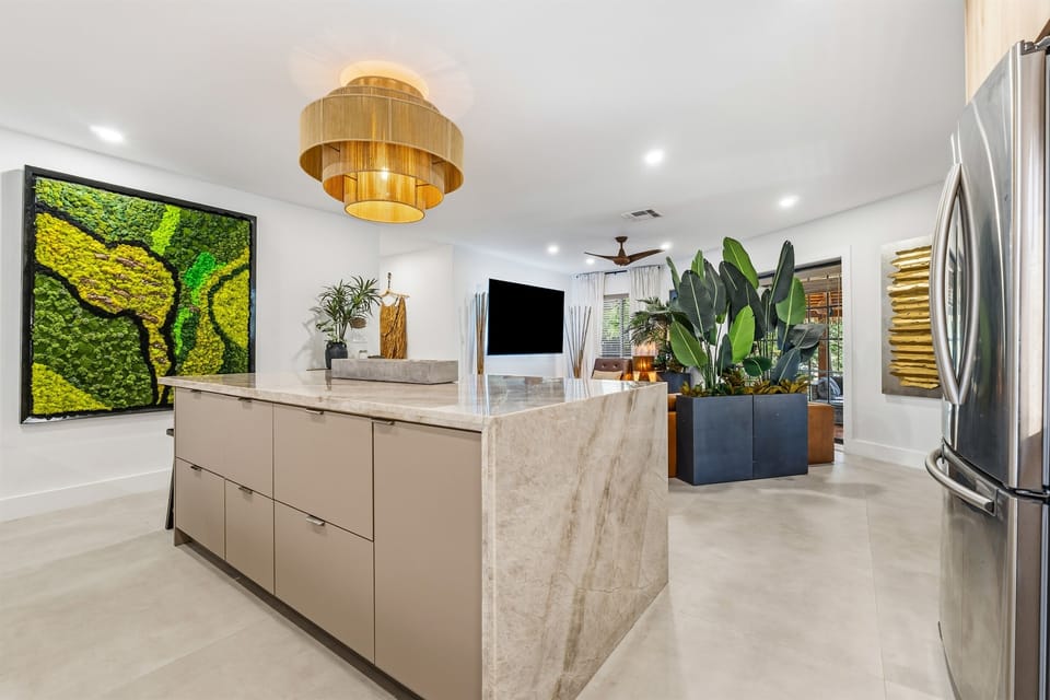 Waterfall quartzite counters, a six-seat island, and smooth cement floors ground this glowing jungle-lux kitchen. Opening into the living room, it keeps the house connected while Tabu Collection moss art and a handwoven chandelier warm the space.