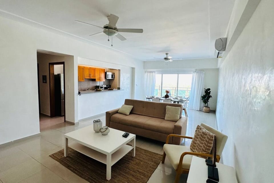 Sala de estar con sofá, con una mesa de madera al centro y Aire acondicionado, al final se aprecia el balcón y la area de cocina